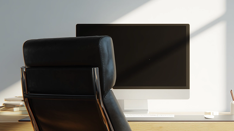 Grand écran d’ordinateur sur un bureau en bois, derrière une chaise en cuir dans une pièce éclairée naturellement.