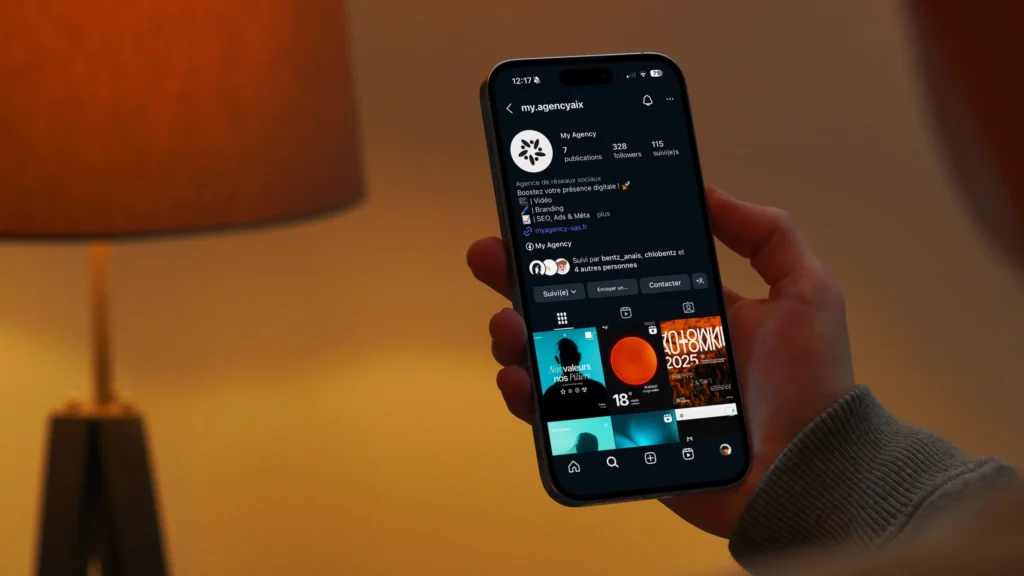Smartphone tenu en main affichant un profil Instagram, avec une lampe allumée en arrière-plan.